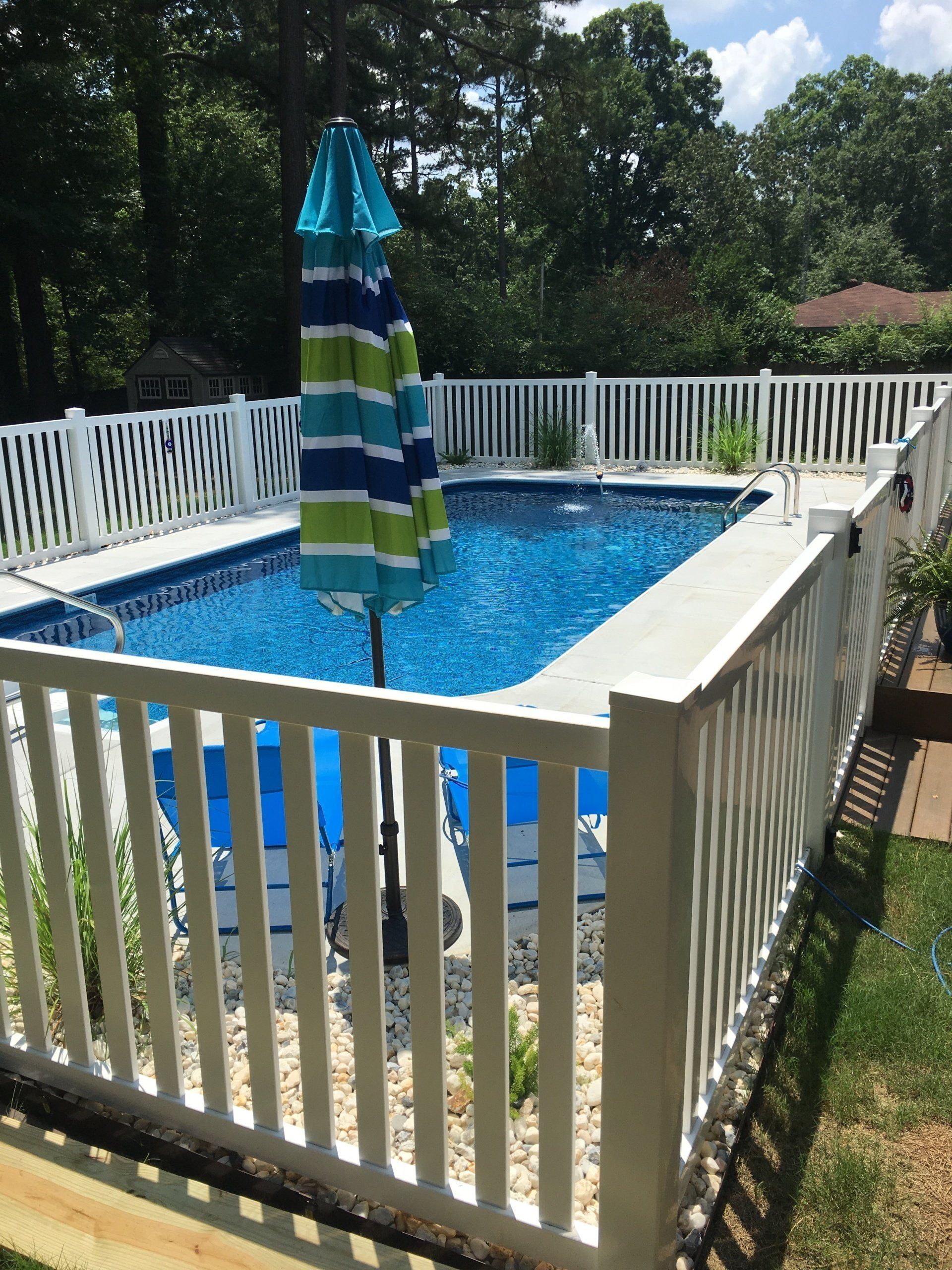 Backyard pool enclosed by a white fence, with a closed striped umbrella in the center.