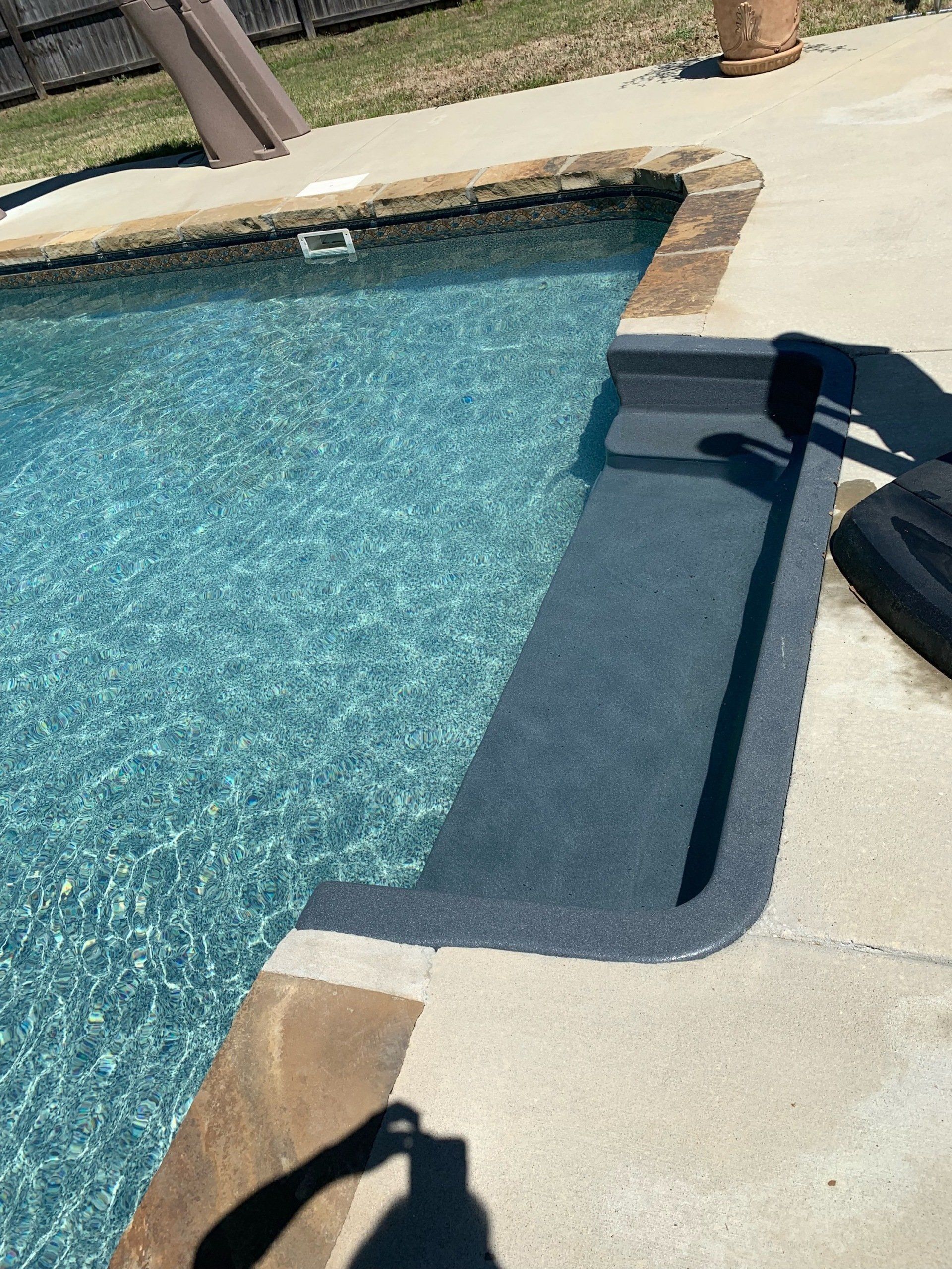 Swimming pool edge with a built-in dark blue step and beige concrete deck in bright sunlight