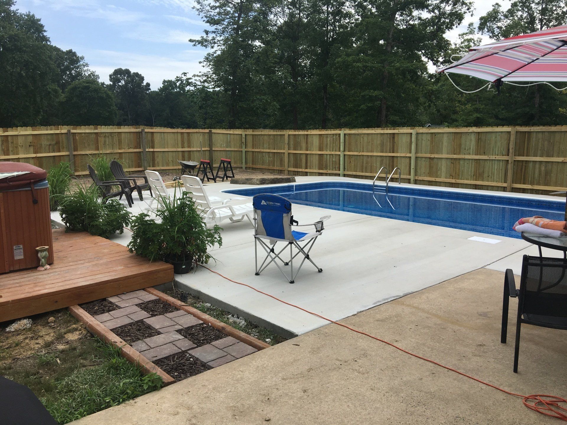 Backyard pool area with patio furniture, blue water, wooden fence, and a folding chair on concrete patio