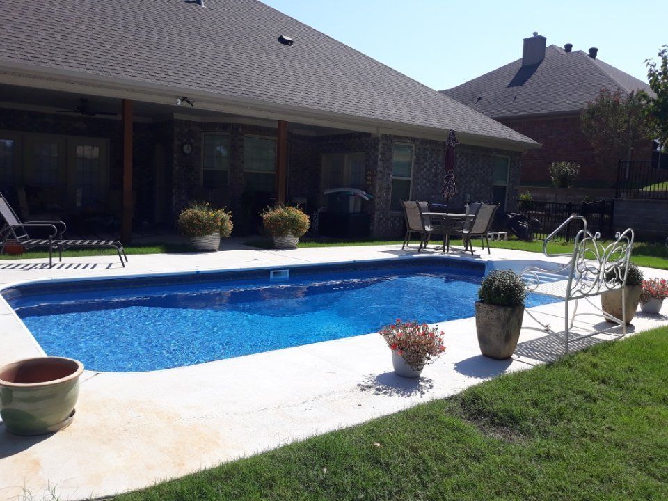 Backyard swimming pool beside a patio and house, with potted plants and a lawn in the foreground