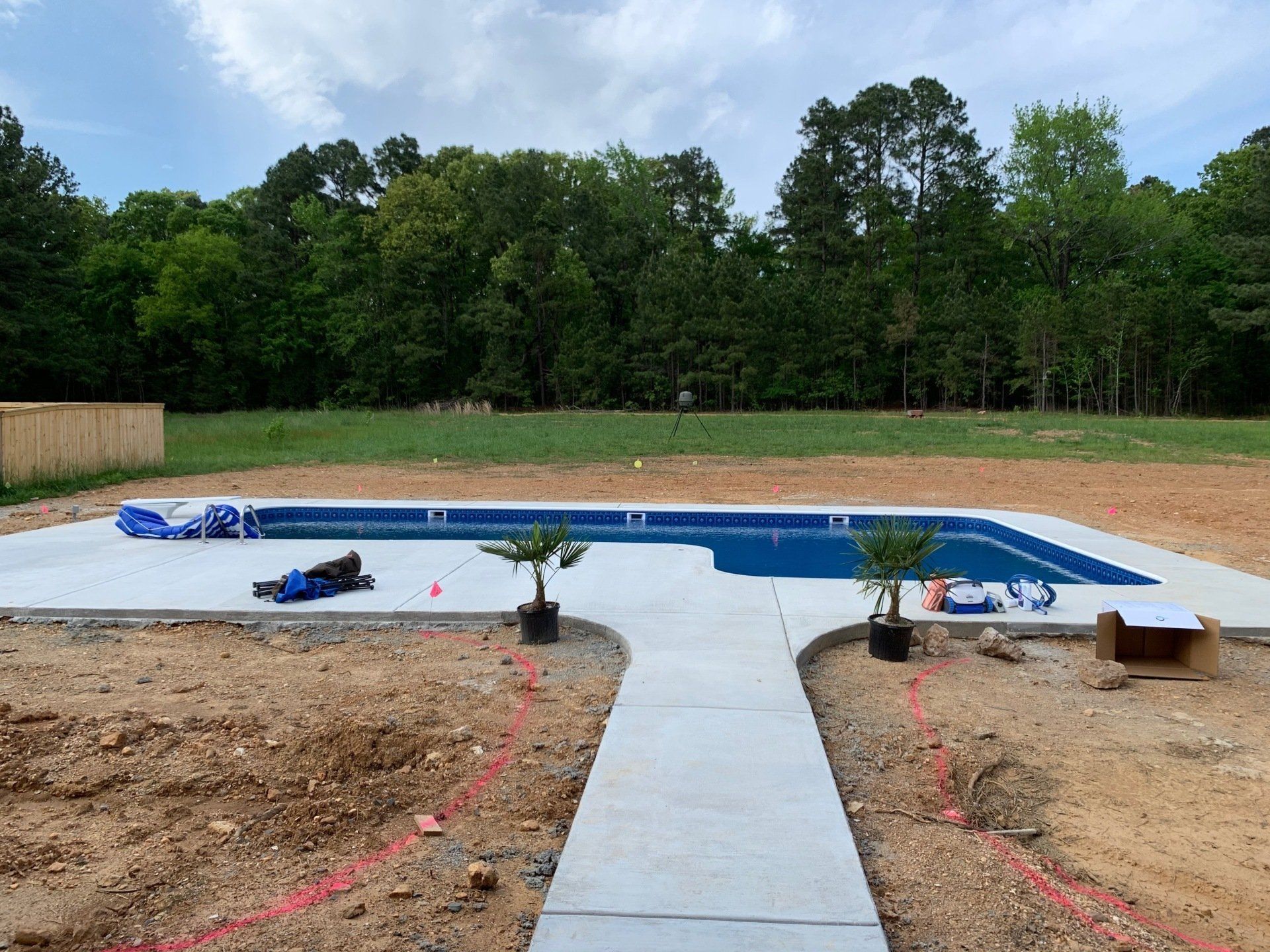 Backyard with a partially filled in-ground pool beside a concrete walkway and trees in the background