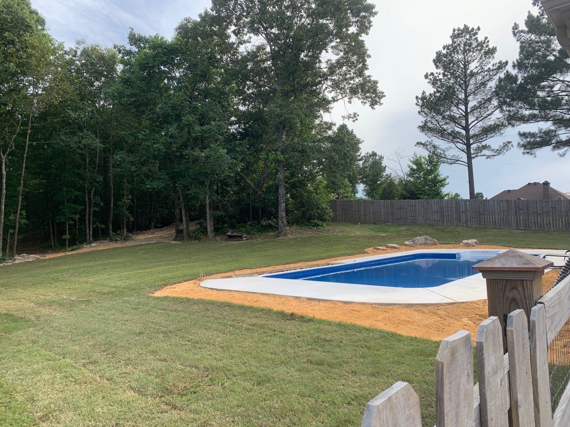 Backyard pool under construction with blue liner, dirt border, grassy yard, and trees beyond a fence