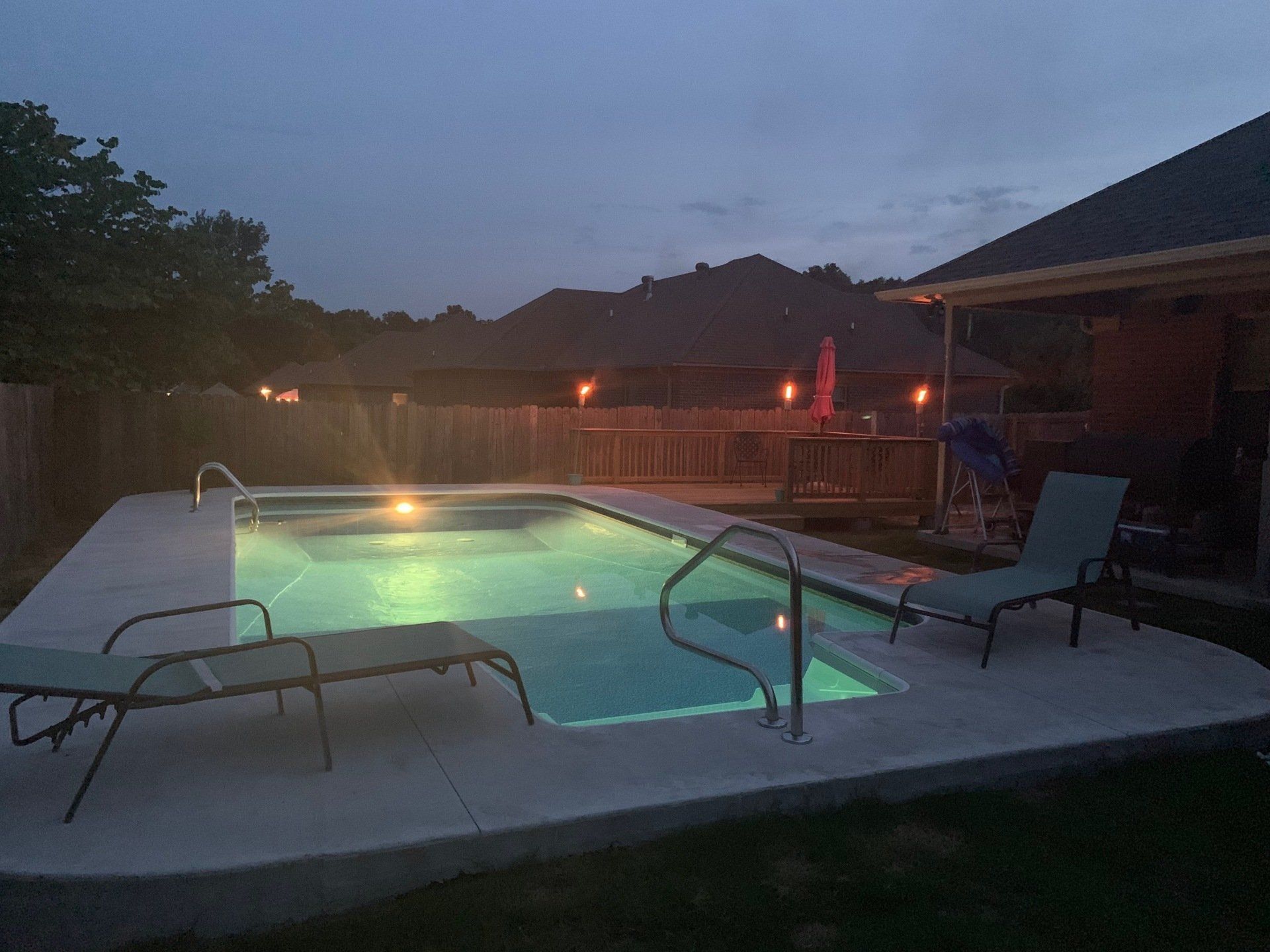 Backyard pool at dusk with glowing underwater lights, lounge chairs, and a patio beside a house