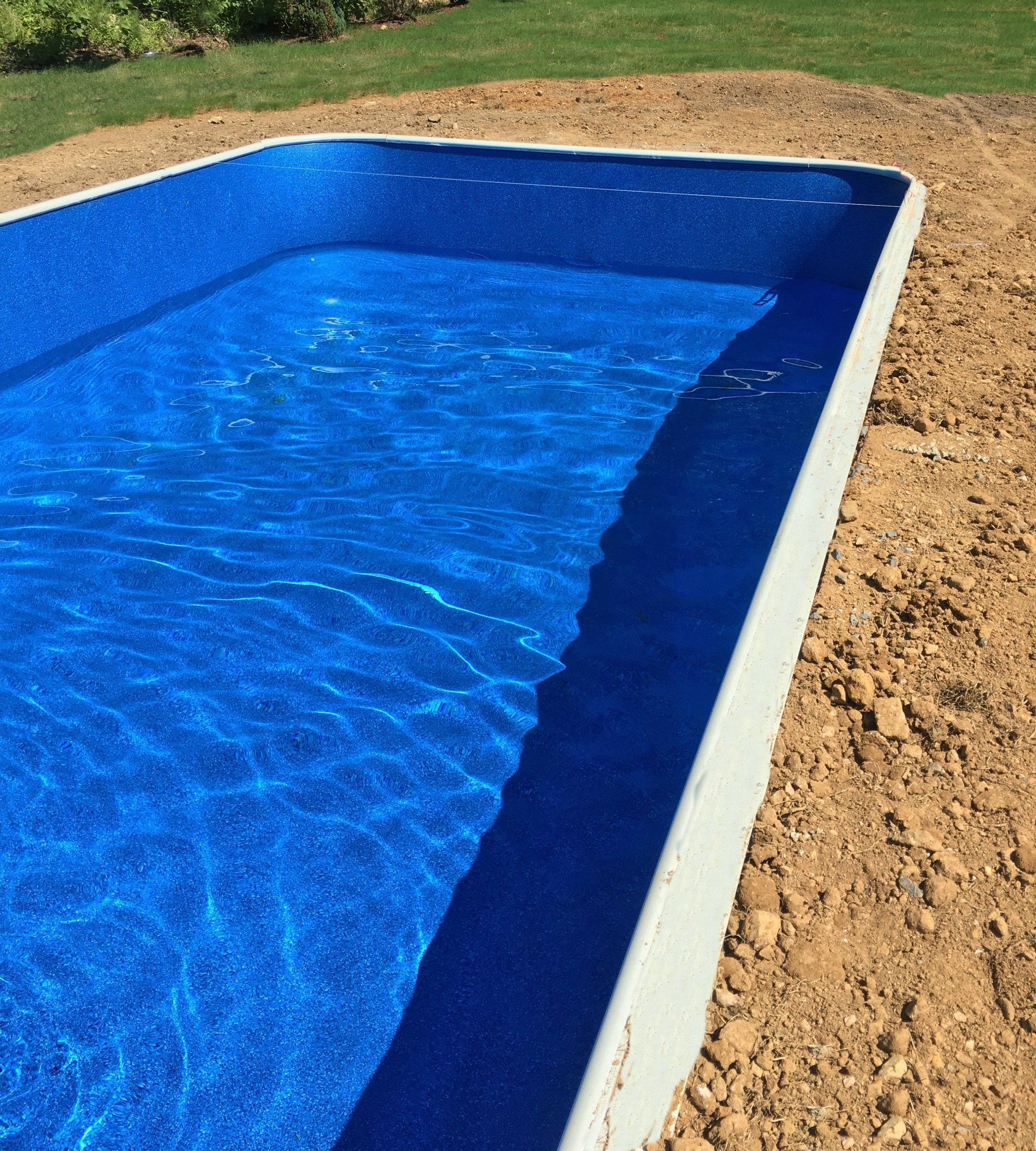 Blue swimming pool filled with water, bordered by white coping and surrounded by dirt and grass