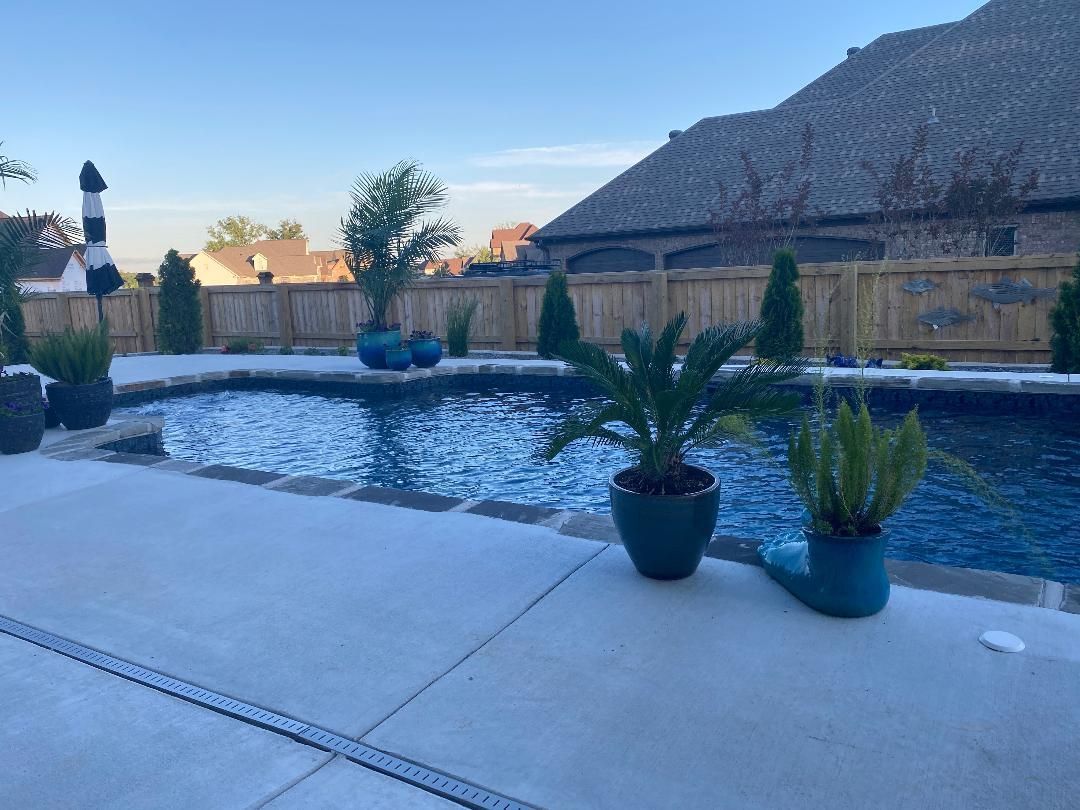 Patio beside a blue-tiled pool with potted plants and a thatched-roof house in the background