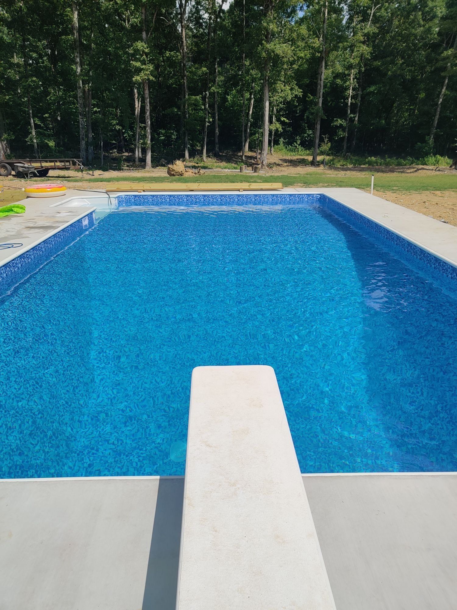 Blue swimming pool with a diving board, surrounded by a wooded backyard.