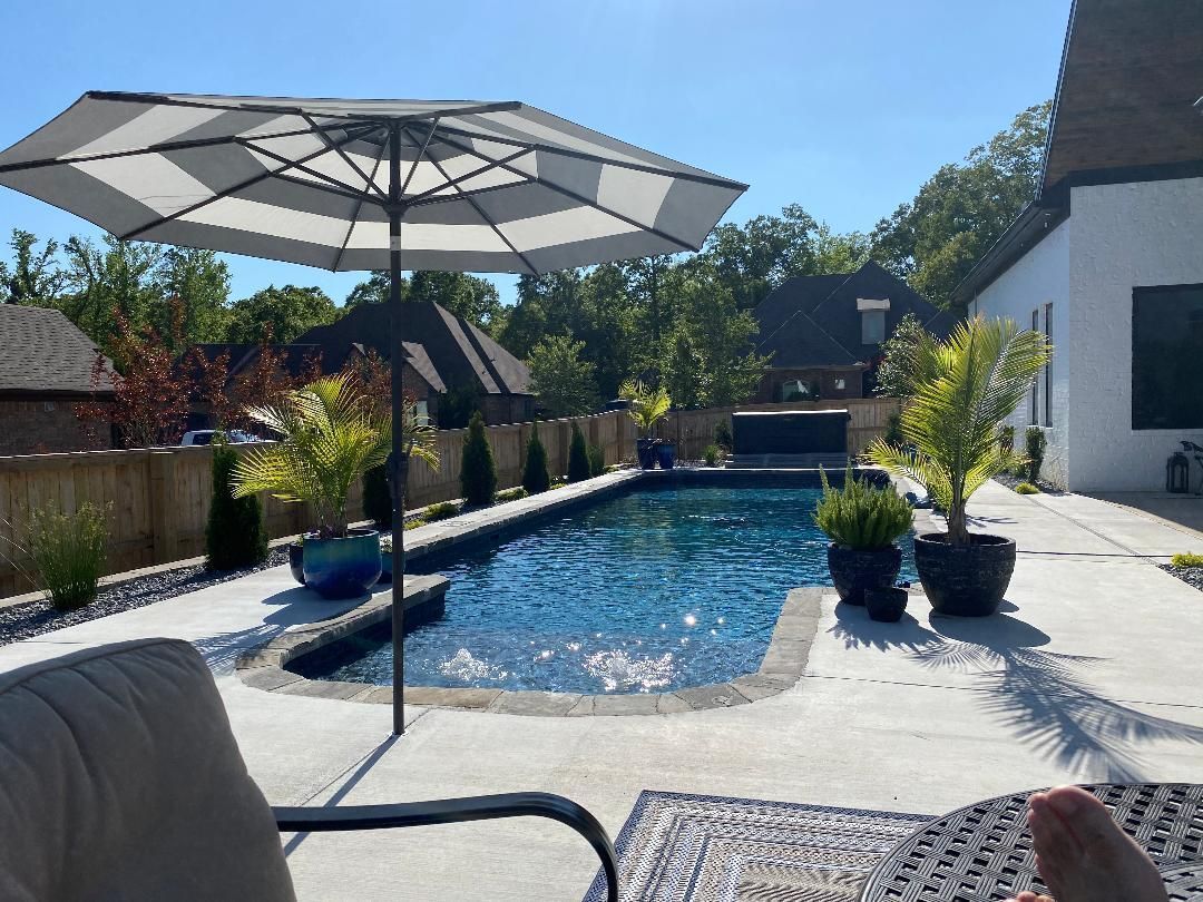 Backyard pool with patio umbrella, lounge chair, and potted plants beside a white house