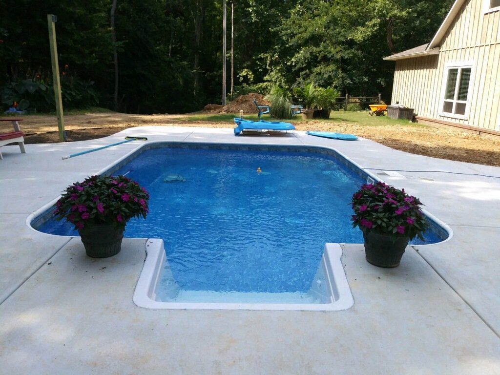Backyard swimming pool with blue water, potted flowers, and a patio beside a house and trees.