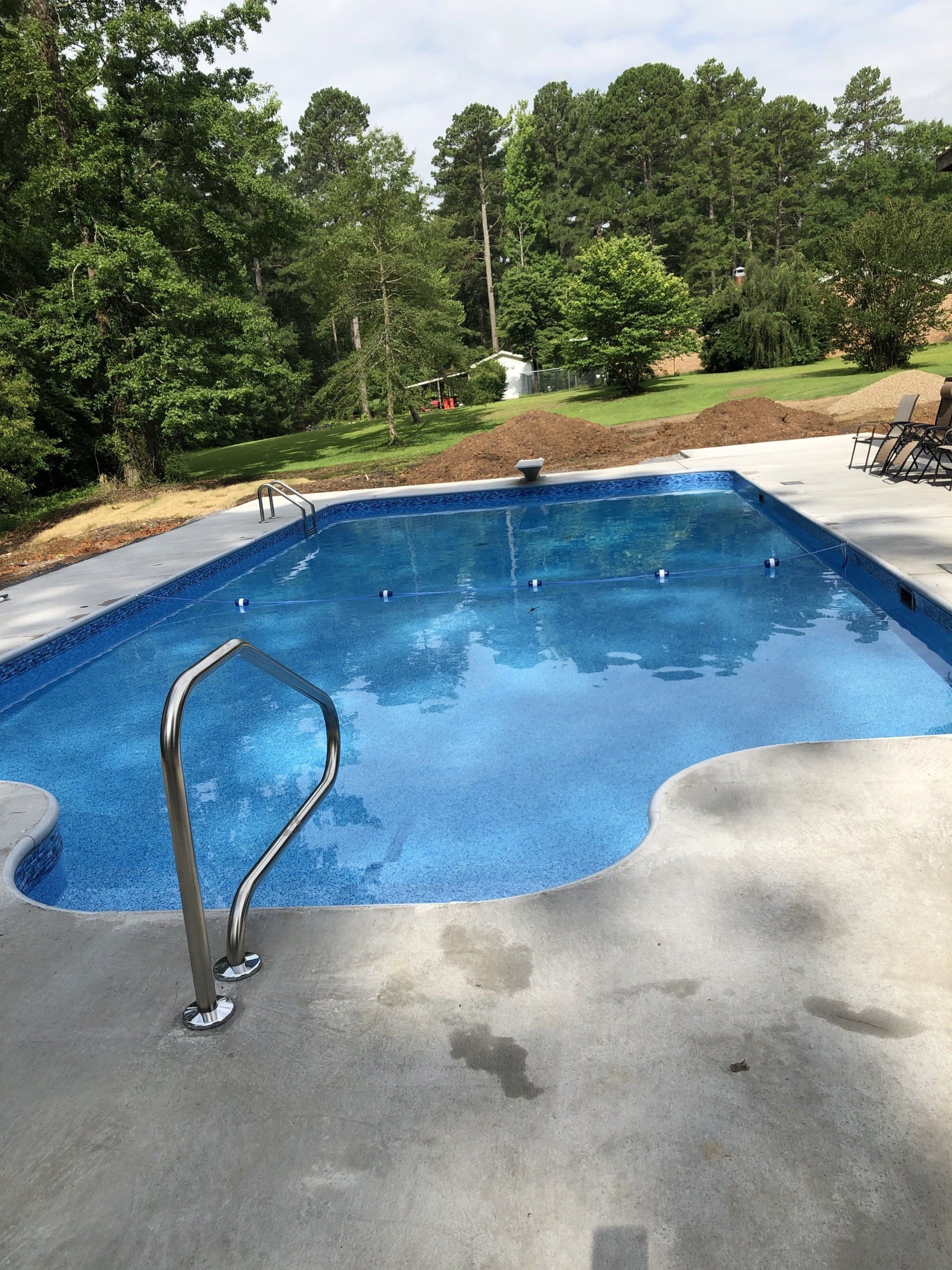 Backyard swimming pool with blue water, concrete deck, and trees in the background