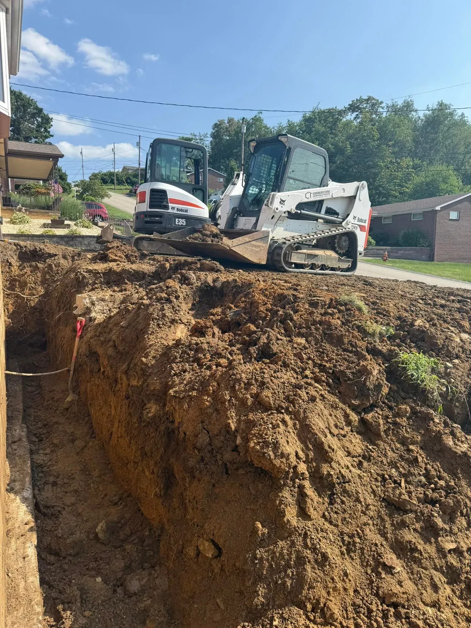 Two Bobcat machines excavating a trench. Earth pile alongside a building. Blue sky and trees in the background.
