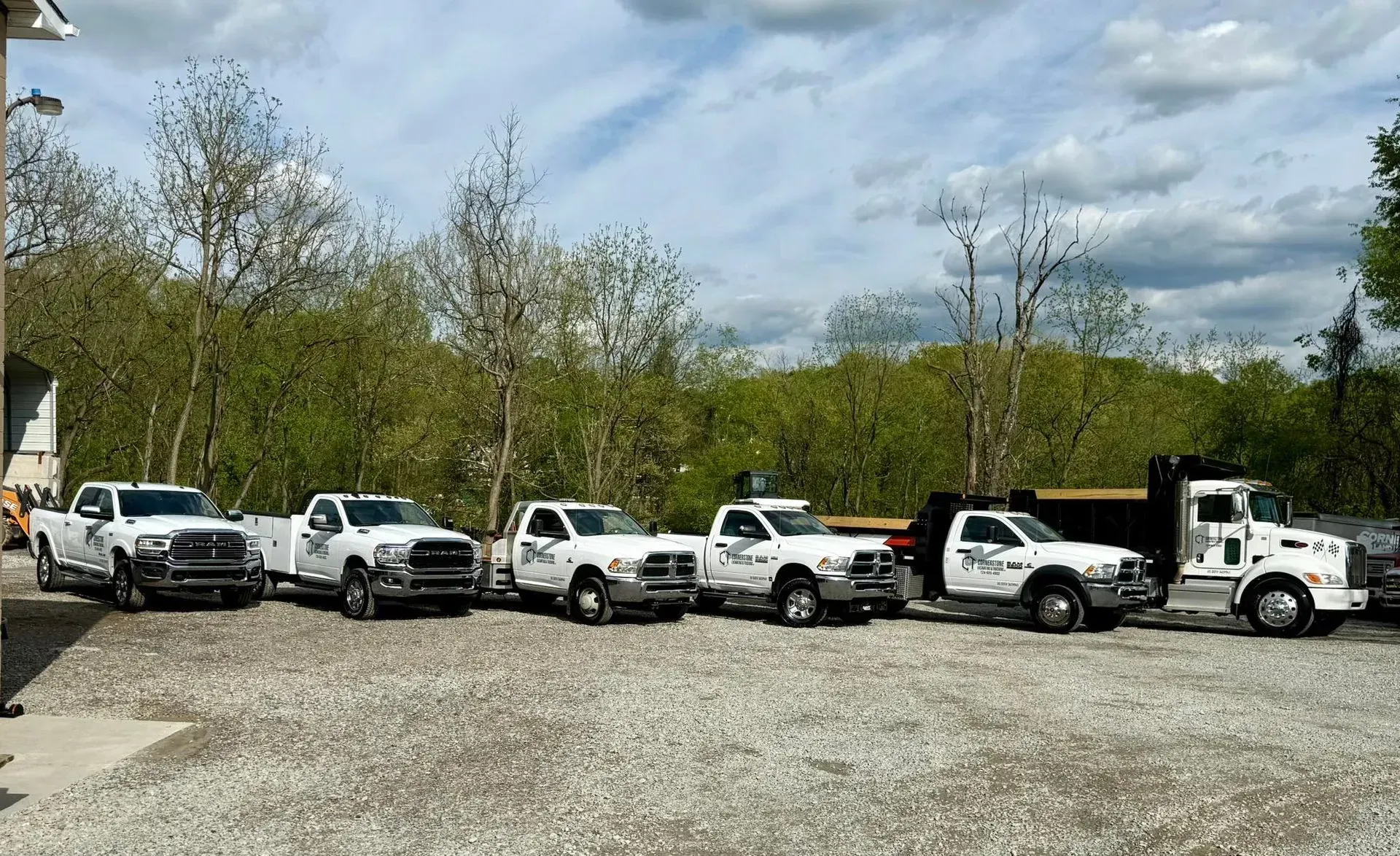 Six white work trucks parked outside, with trees in the background.