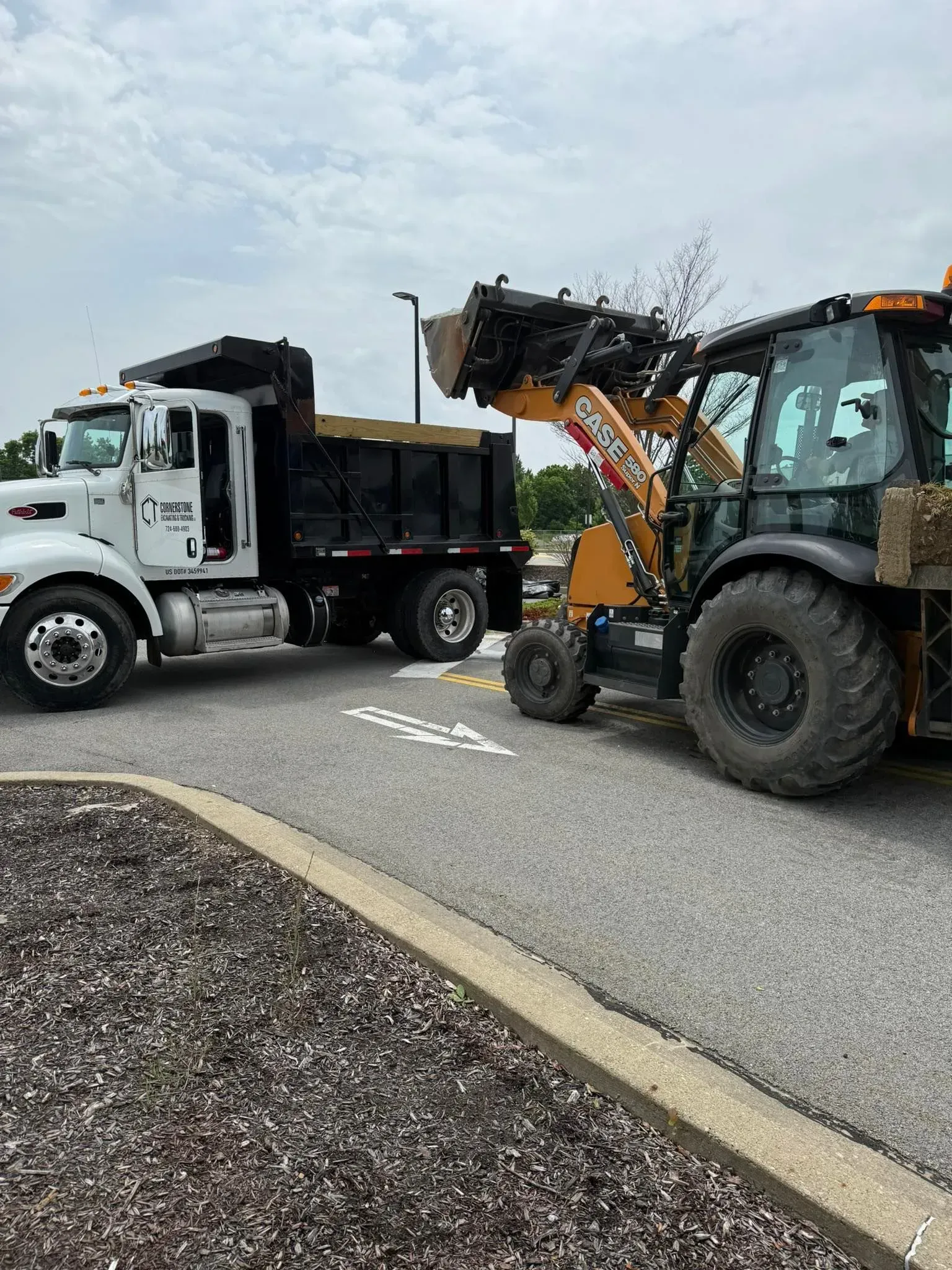 A backhoe loading debris into a dump truck on a paved surface. Cloudy sky.