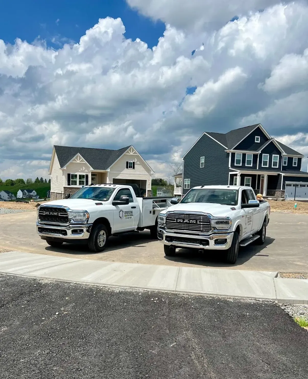 Two white RAM trucks parked in front of new houses on a sunny day with a cloudy sky.