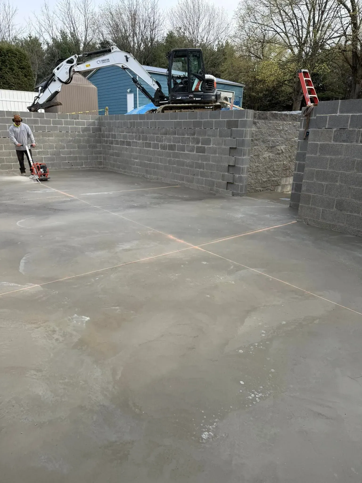 Man smoothing concrete, excavator in background, construction site.