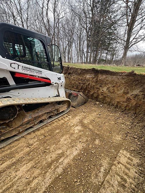 White Bobcat excavator digging a trench in a dirt field. Trees in the background.