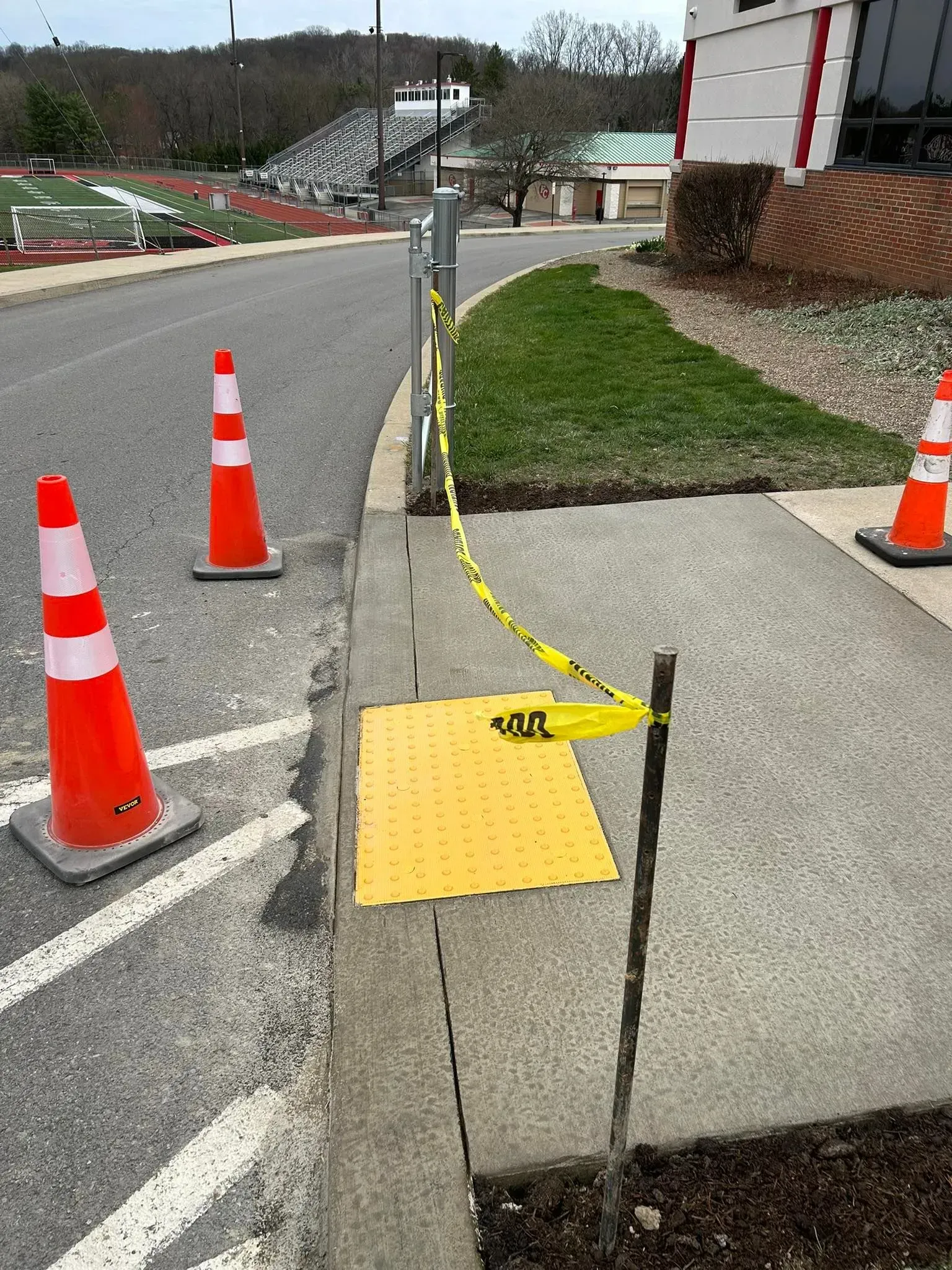 Orange traffic cones and caution tape block a sidewalk ramp, potentially impeding pedestrian access.