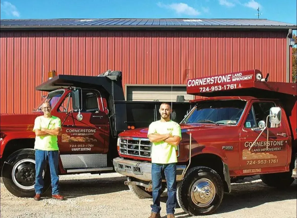 Two men stand with arms crossed in front of red dump trucks, outside a red building, under a blue sky.