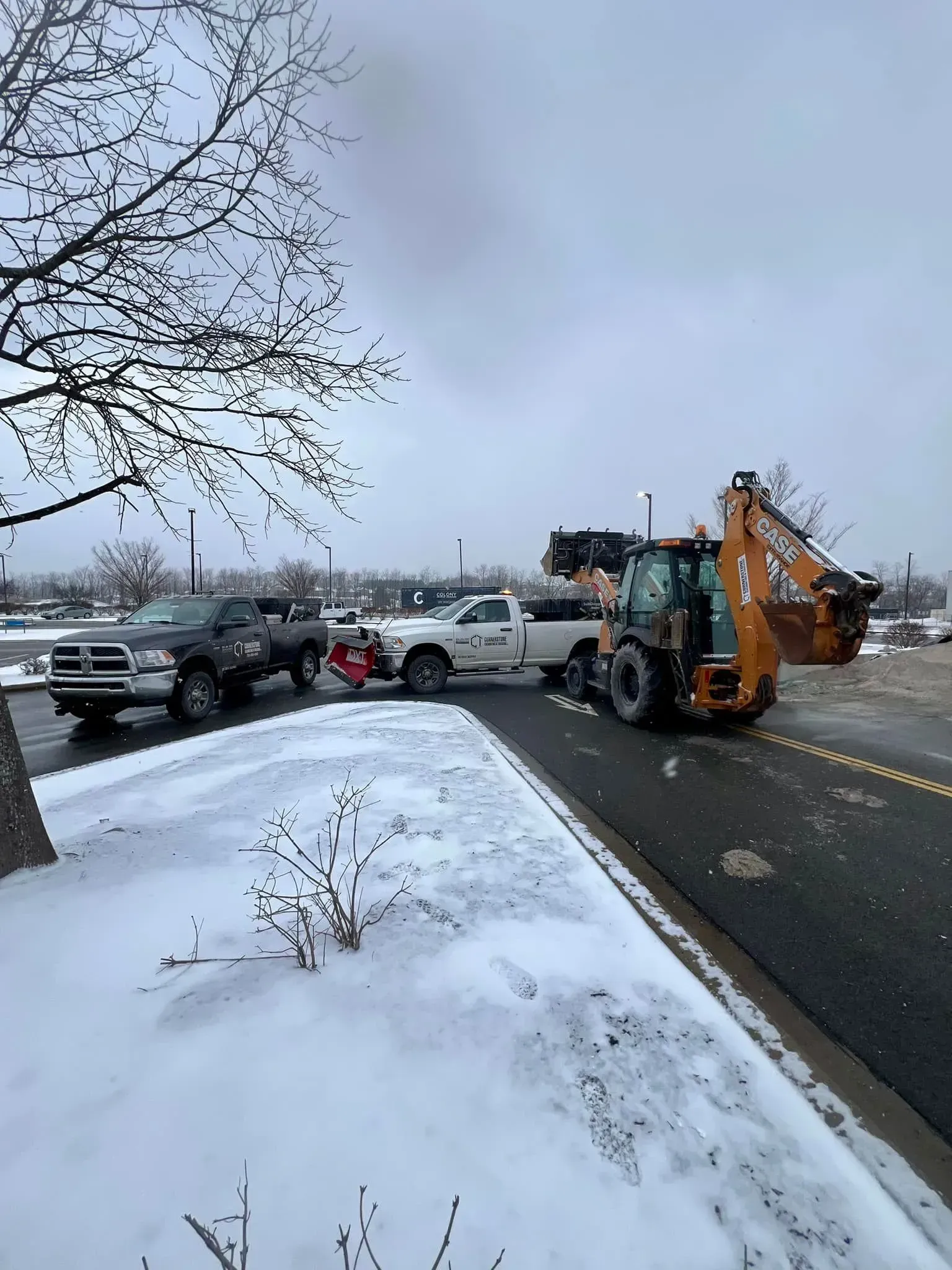 Snow removal equipment clearing a parking lot: pickup trucks with plows and a backhoe in a winter setting.
