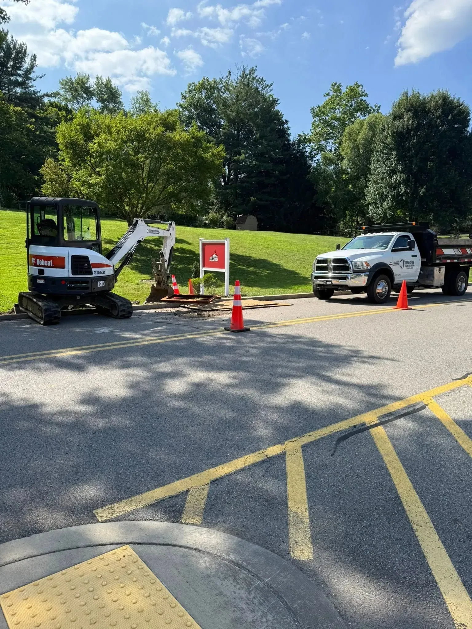 Bobcat excavator and work truck at road construction site, with orange cones and sign.