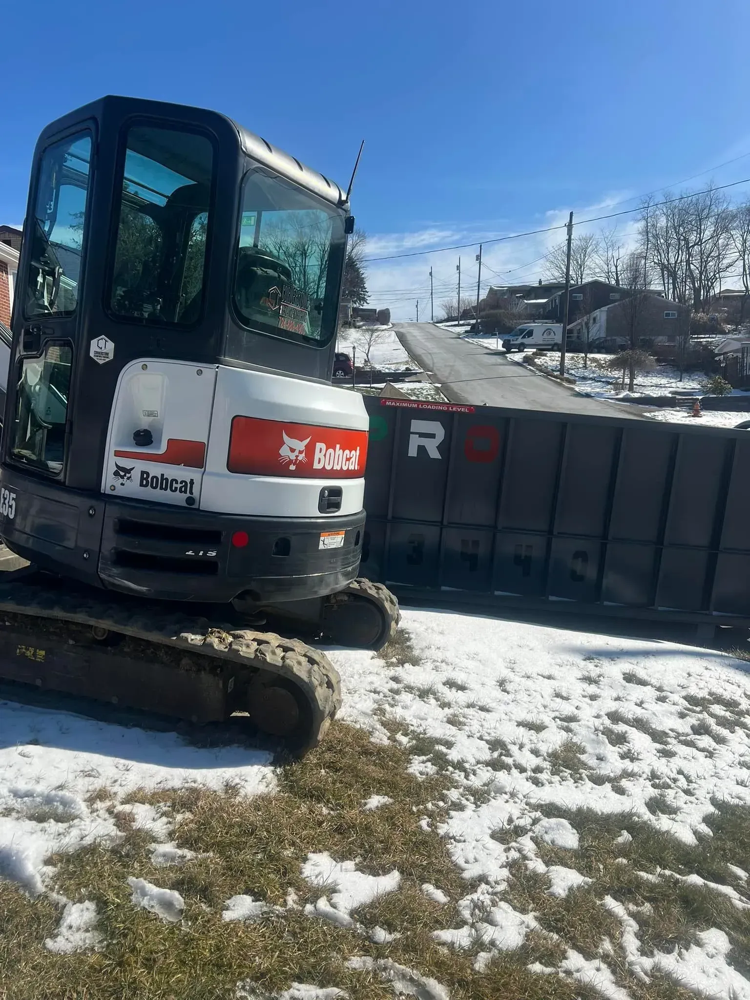 Bobcat excavator next to large gray blocks in a snowy, sunny outdoor setting.