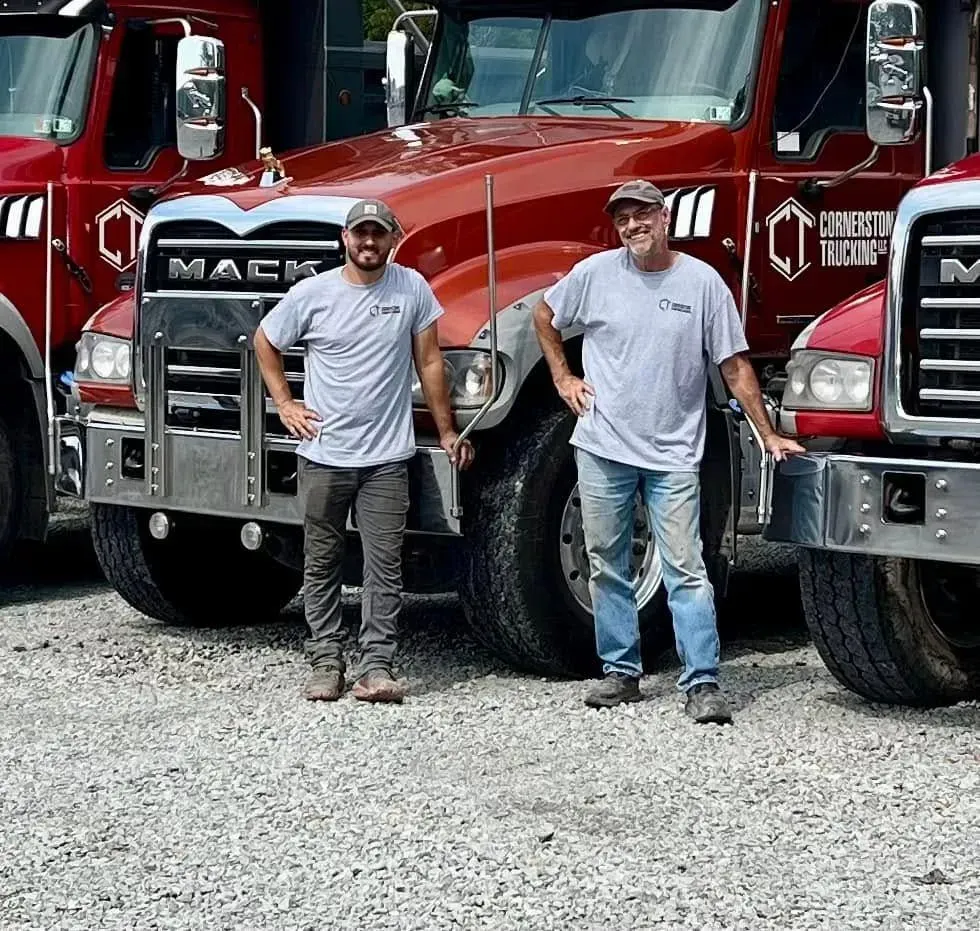 Two men in front of red Mack trucks; one man wearing camo, the other in jeans, smiling.