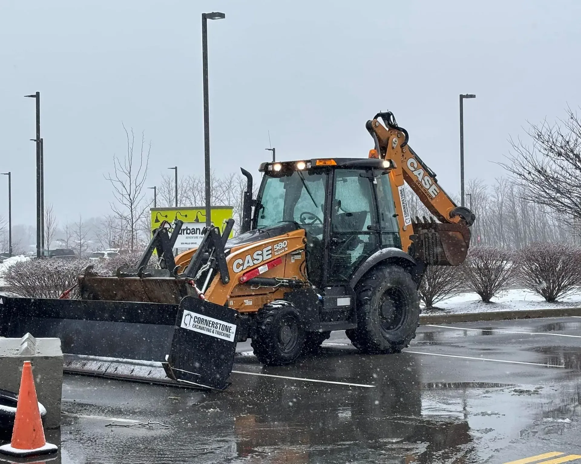 A backhoe with a snowplow clearing a wet parking lot on a snowy day.