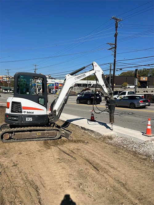Bobcat excavator breaking concrete curb along a road with parked cars and buildings.