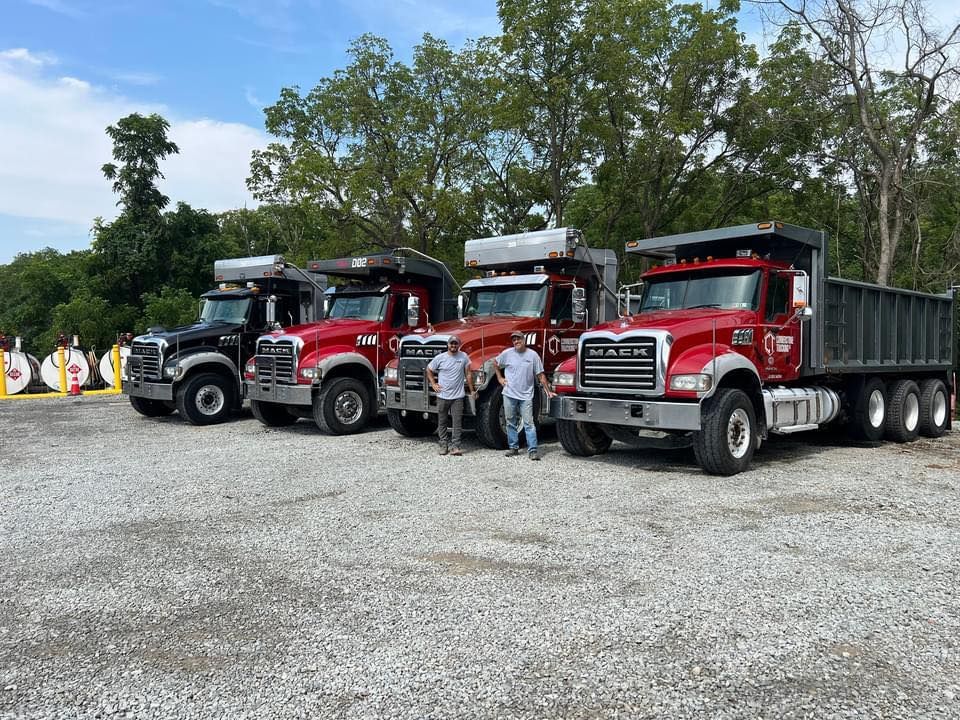 Four dump trucks of varying colors parked outdoors, with two men posing in front.