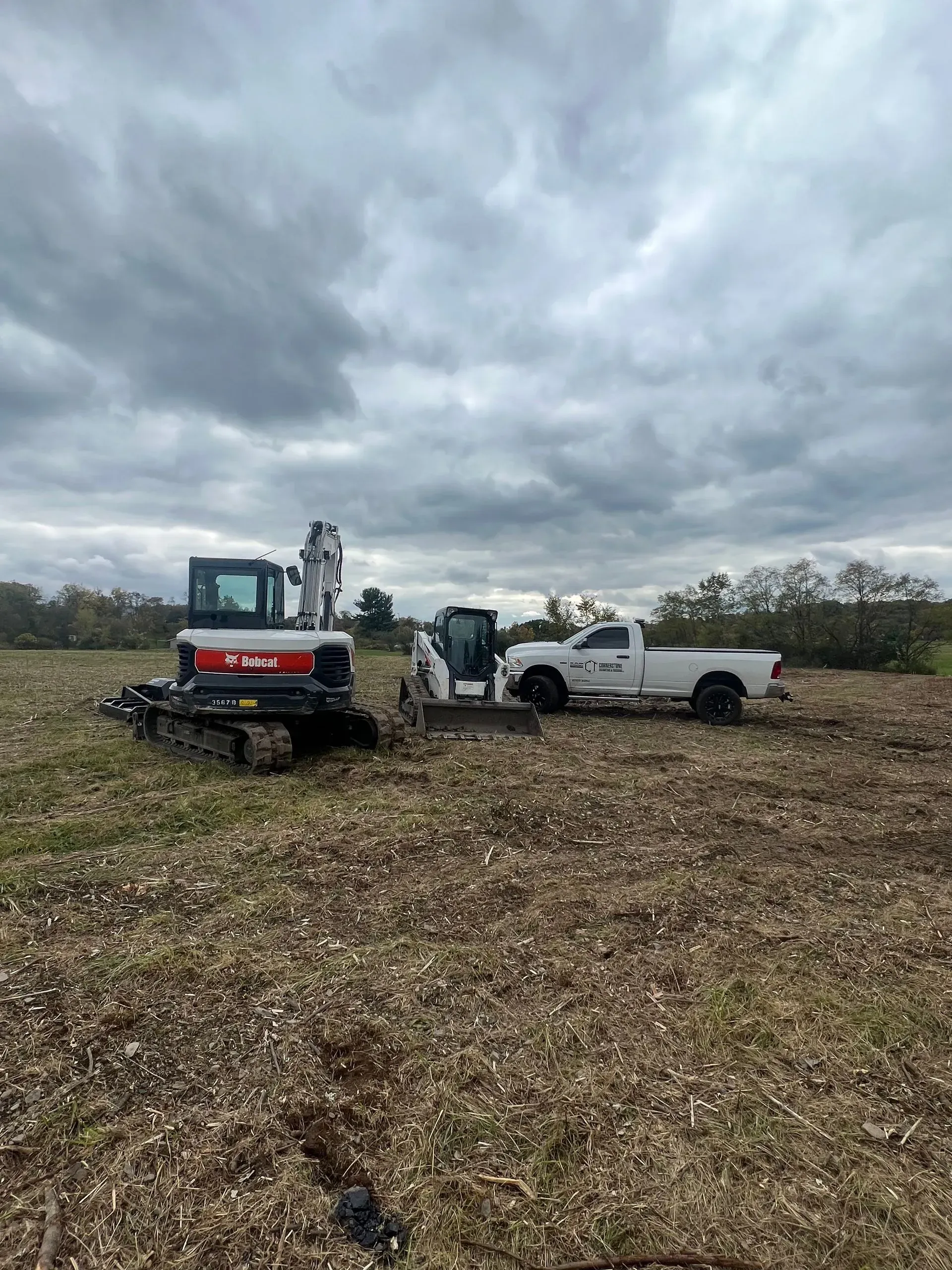 Bobcat track loader, truck, and worker clearing land under a cloudy sky.