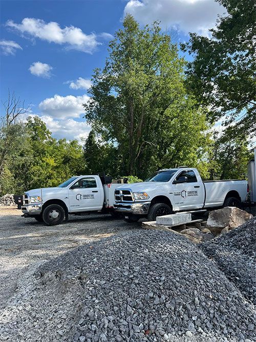 Two white work trucks parked near a pile of gravel under a blue sky.