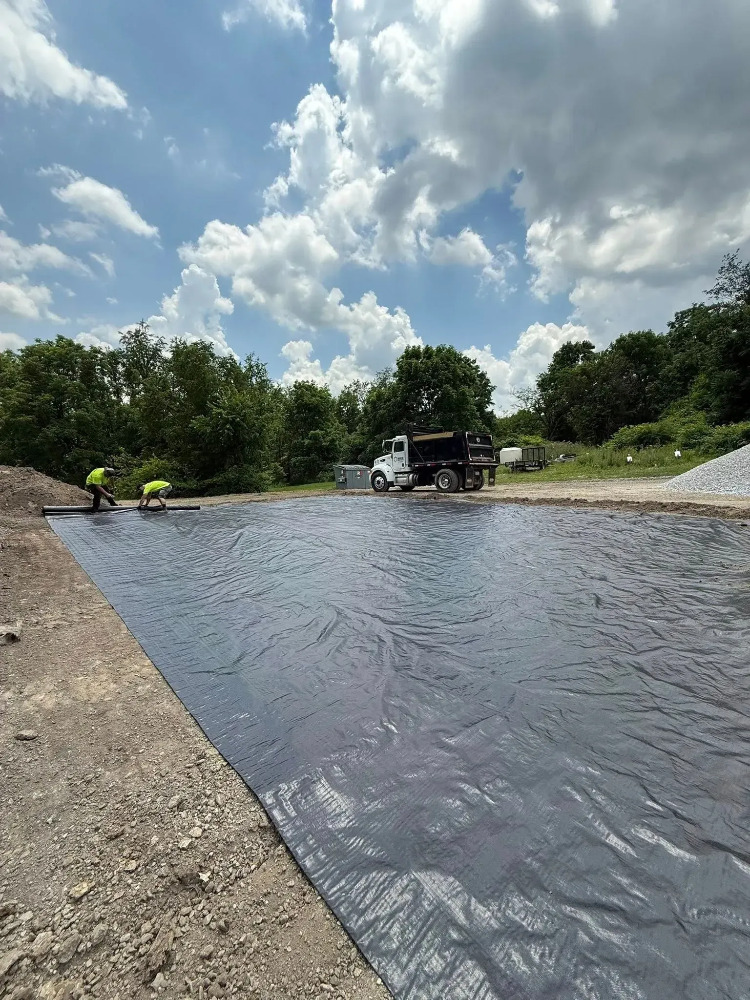 Two workers installing black fabric at a construction site. A truck is nearby, and the sky is cloudy.