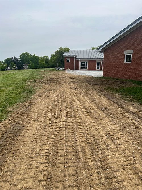 Dirt path with tire tracks leading to brick buildings with metal roofs, on a cloudy day.