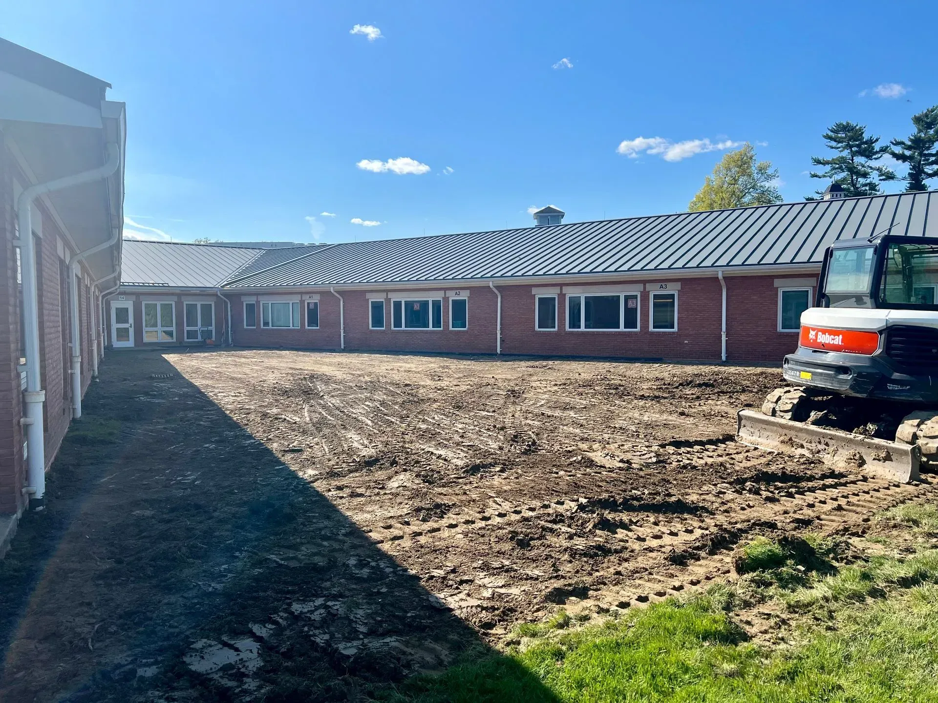 A red brick building under construction with a small excavator on a dirt area under a blue sky.