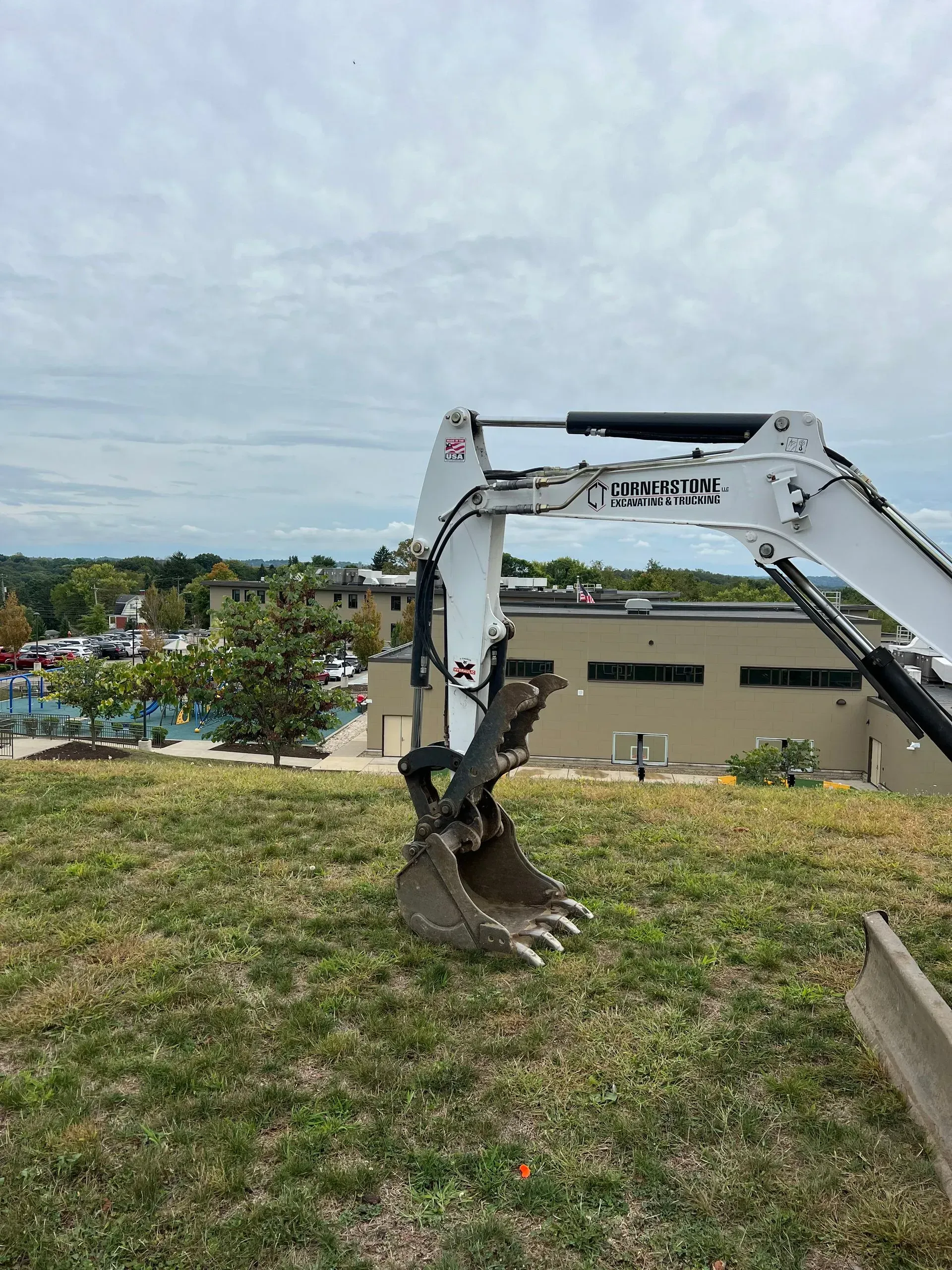 White excavator bucket on a grassy hill, with a building and trees in the background under a cloudy sky.