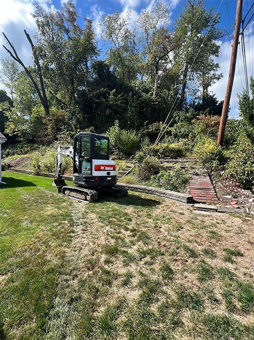 Bobcat excavator on a grassy slope near a retaining wall and trees.