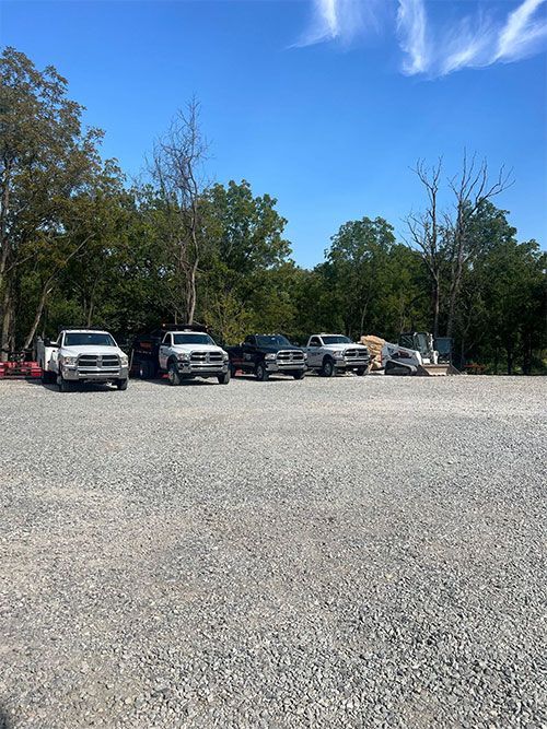 Four trucks parked on a gravel lot under a blue sky, trees in background.