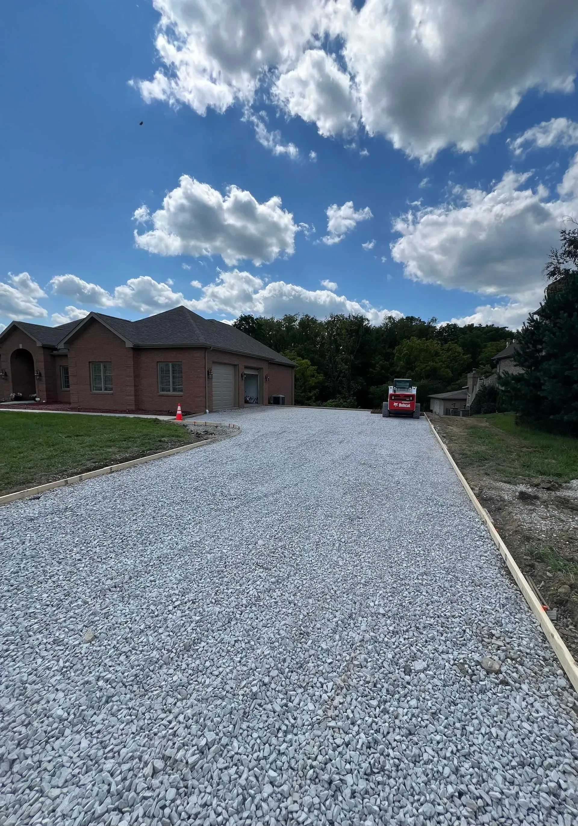 Gravel driveway leading to a brick house on a sunny day; a red skid steer is parked at the top.