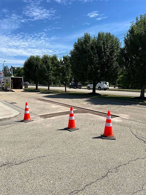 Three orange traffic cones block a section of cracked asphalt. Trees and a blue sky are in the background.