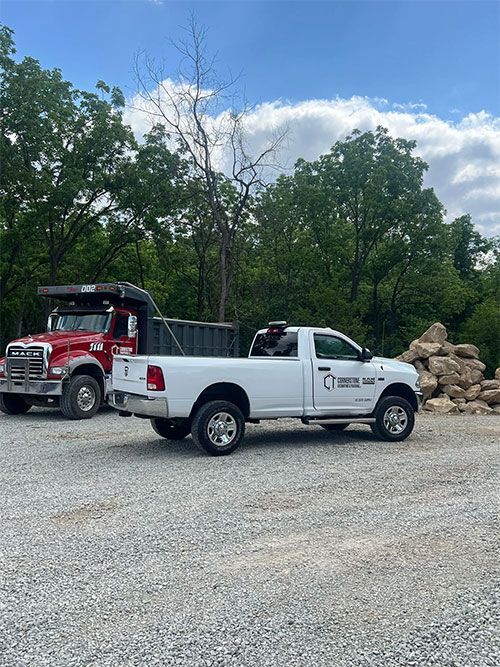 White pickup truck with portable toilets parked near red dump truck in a gravel lot; trees in background.