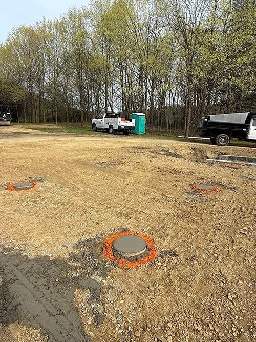 Construction site with concrete pads marked by orange paint; trucks, porta-potty, trees in background.