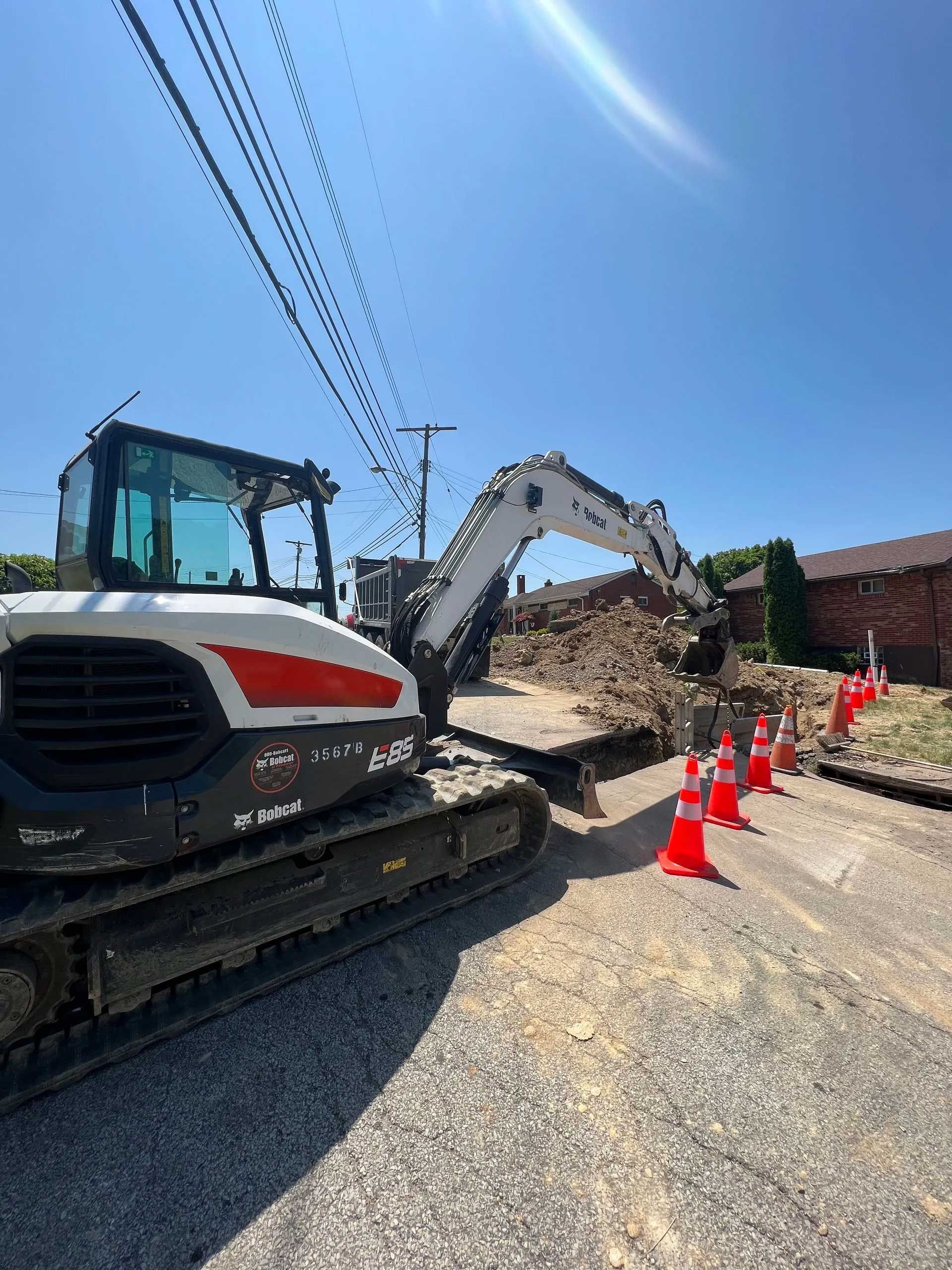 Bobcat excavator digging in a road, orange cones line the work area, under a blue sky.