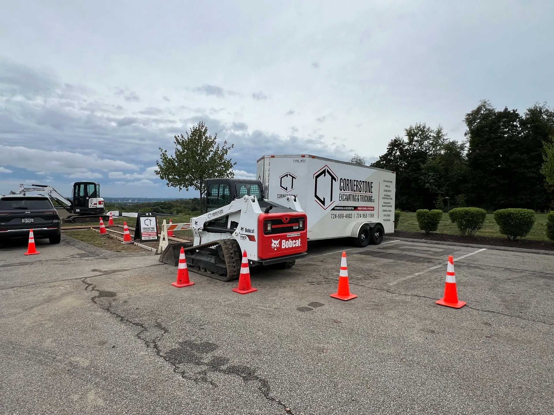 Bobcat construction equipment and trailer on asphalt with safety cones, cloudy sky.