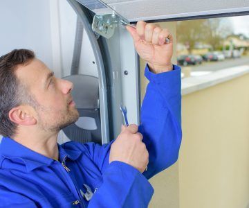 Man in blue overalls repairs a garage door, using a wrench. Inside a garage, in front of an open window.