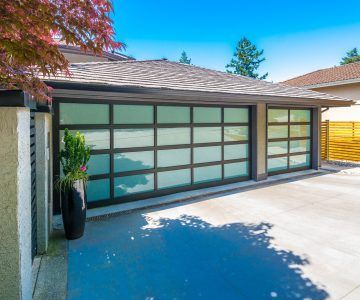 Modern garage with glass doors, black frames, and concrete driveway. Sunny day, lush landscaping.