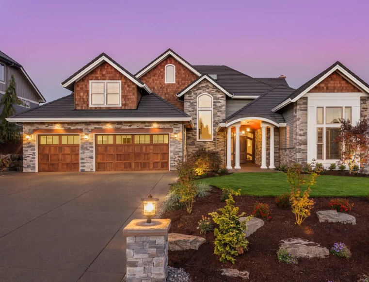 Luxurious two-story home with stone and wood exterior, a driveway, and manicured landscaping at dusk.
