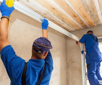 Two workers installing molding on a wall, one using a drill, in a room with a wooden ceiling.