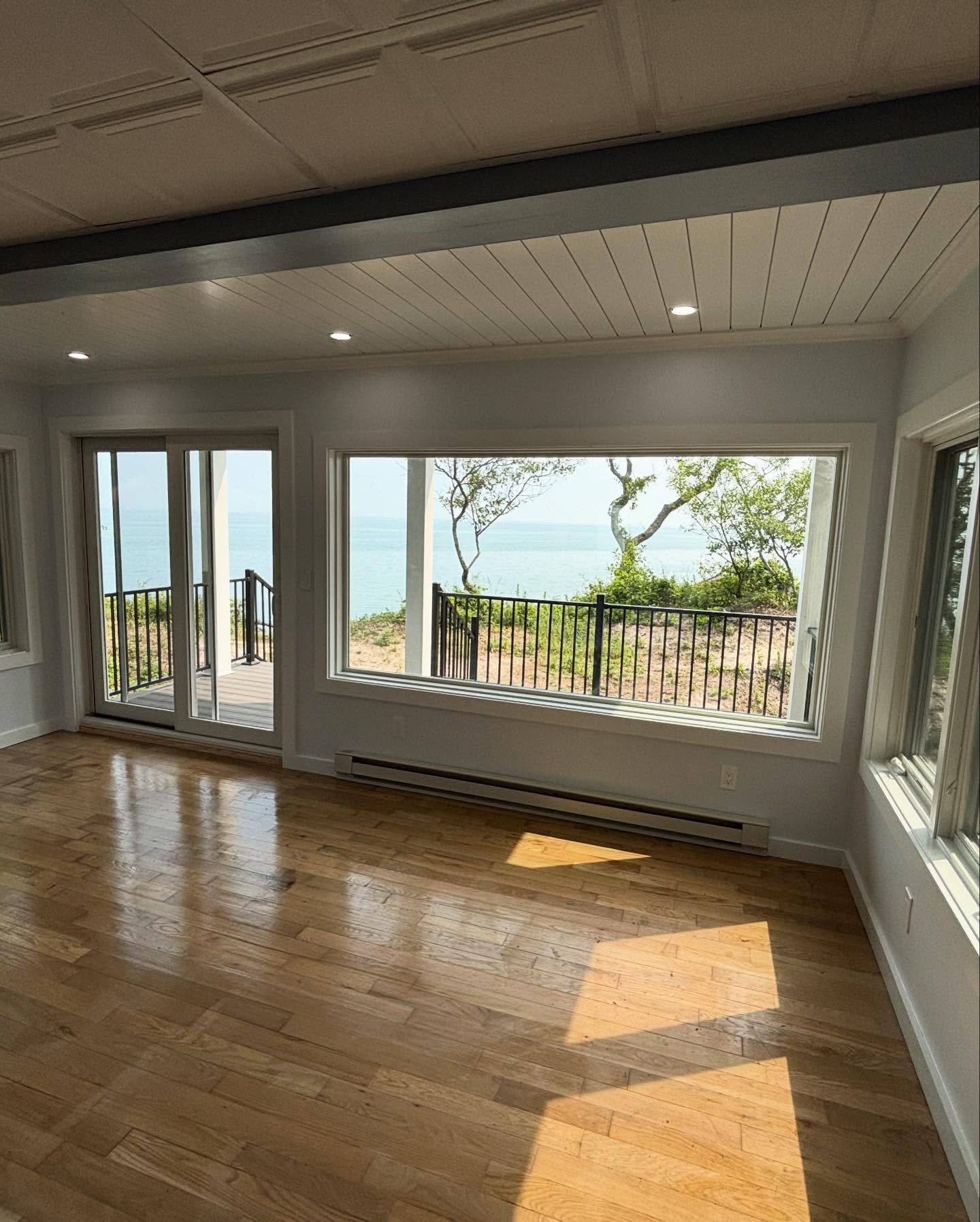 Empty sunlit room with hardwood floors, large windows, and ocean-view balcony doors