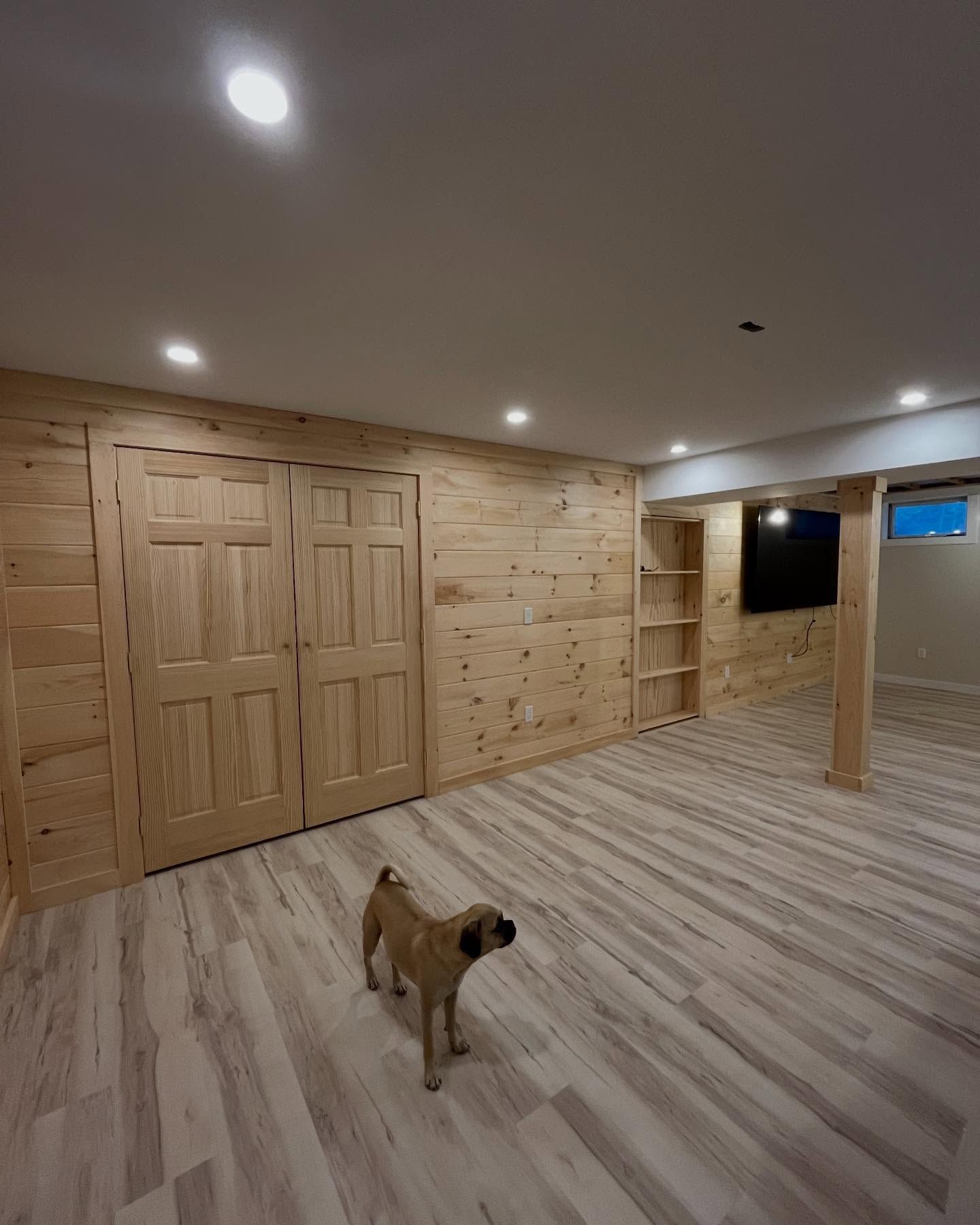Spacious unfinished basement with wood-paneled walls, recessed lights, and a small dog standing on wood flooring.