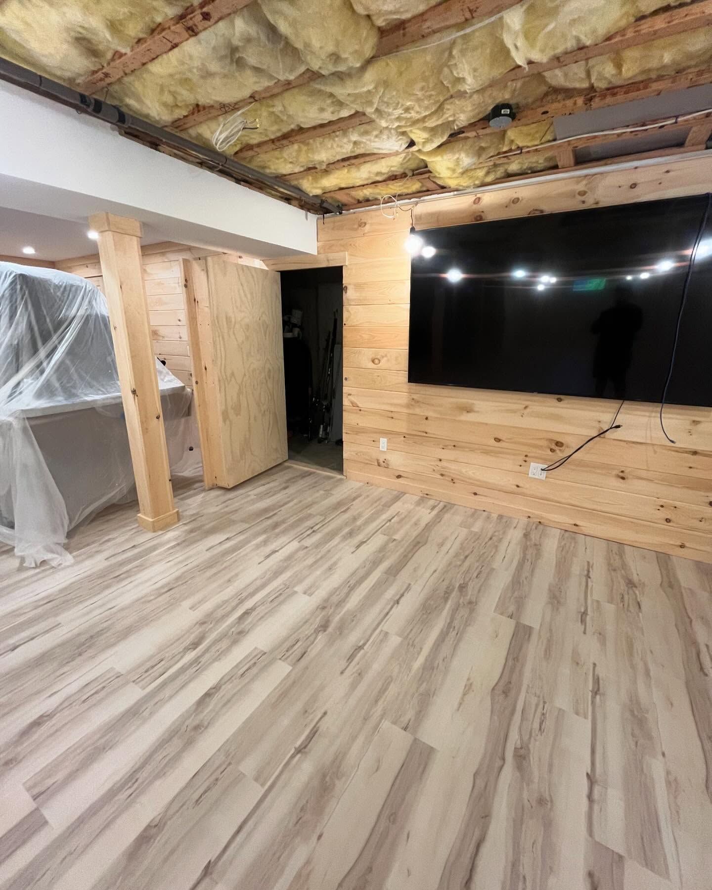 Basement room under renovation with wood-look floor, exposed insulation ceiling, and large wall-mounted TV area