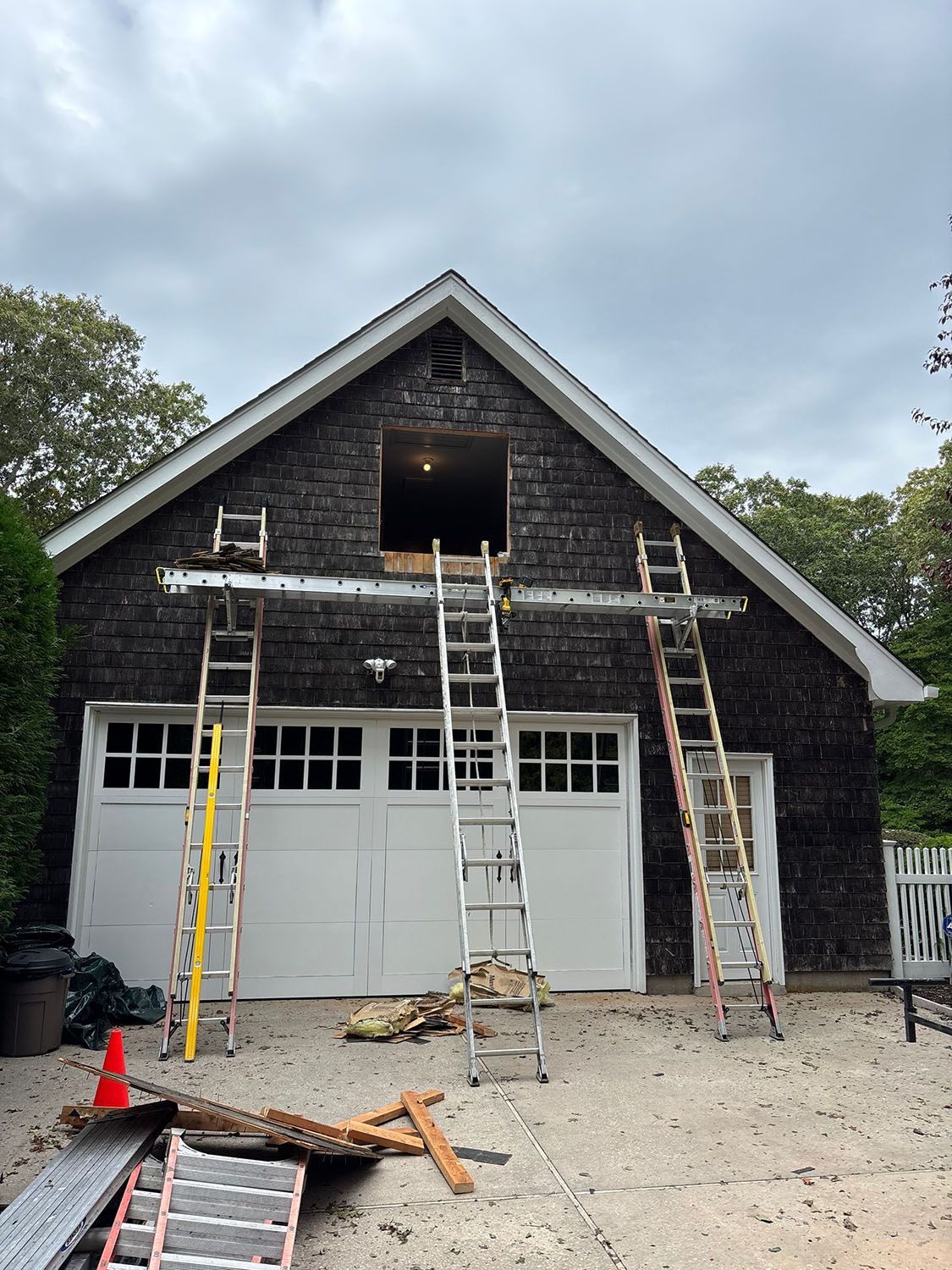 Two-story house with dark siding under renovation, ladders leaning on the front and debris on the driveway.
